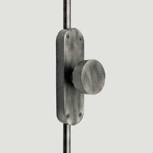 Op de afbeelding: Een donkergrijze metalen deurknop gemonteerd op een ovaalvormige achterplaat met een geborstelde afwerking. Het geheel is bevestigd aan een verticale stang en vastgezet met vier zichtbare schroeven aan de boven- en onderkant van de plaat, met een eenvoudige cilindrische knop.
