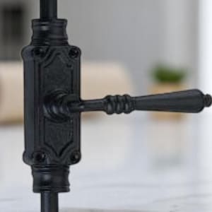 Op de afbeelding: Close-up van een zwarte, sierlijke deurklink. De klink heeft een gedetailleerd ontwerp met een hendel en een decoratieve achterplaat. De klink is gemaakt van metaal en heeft een matte afwerking. De achtergrond is wazig, wat wijst op een interieur.