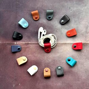 Op de afbeelding: Een verzameling kleurrijke leren kabelorganizers, gerangschikt in een cirkel rond een paar witte oortelefoons. De organizers zijn in verschillende kleuren, waaronder zwart, bruin, rood en blauw, elk met een drukknoopsluiting.