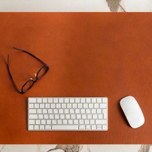 Könnte beinhalten: Eine braune Leder-Schreibtischunterlage mit einer weißen kabellosen Tastatur und Maus. Ein Paar schwarze Brillen liegt auf der Unterlage.