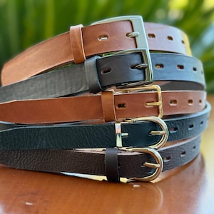 Op de afbeelding: Een stapel leren riemen in verschillende kleuren, waaronder bruin, zwart en grijs. Elke riem heeft een metalen gesp. De riemen zijn netjes opgestapeld en tonen hun verschillende tinten en texturen.