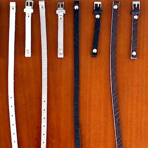 Peut inclure: Un ensemble de huit fines lanières en cuir blanc, beige, noir et marron. Quatre lanières sont dotées de boucles en métal argenté et de rivets dorés, tandis que les quatre autres sont de longues bandes simples avec des trous de réglage.