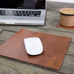 May include: A brown leather mouse pad with a white wireless mouse on top. The mouse pad is on a wooden table with a laptop, a yellow sticky note, a pen, and a coffee cup on a coaster.
