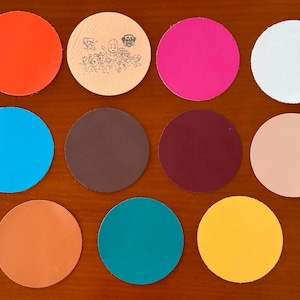 May include: Eleven circular coasters in various solid colors including orange, bright pink, white, blue, brown, dark red, beige, peach, teal, and yellow. One beige coaster features a black line art graphic of Paw Patrol characters.