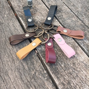 Puede incluir: Seis llaveros de cuero con anillos de metal en varios colores, incluyendo marrón, negro, rosa, amarillo y burdeos. Los llaveros están dispuestos sobre una superficie de madera.