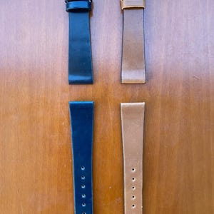 Könnte beinhalten: Vier Leder-Armbänder in Schwarz und Braun. Zwei Armbänder haben silberne Schnallen und sind befestigt. Die anderen beiden Armbänder sind unbefestigt, mit mehreren Löchern zur Anpassung. Die Armbänder werden auf einer Holzoberfläche präsentiert.