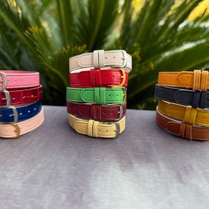 Könnte beinhalten: Gestapelte farbige Lederarmbänder in Rosa, Rot, Blau und Beige. Weitere Stapel umfassen grüne, weinrote und gelbe Armbänder. Der letzte Stapel hat orange, schwarze, gelbe und braune Armbänder. Jedes Armband hat eine silberne Schnalle.