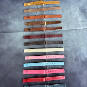 Może przedstawiać: Kolekcja skórzanych pasków w różnych kolorach, w tym beżowym, brązowym, pomarańczowym, różowym, niebieskim, czerwonym i czarnym. Każdy pasek posiada zapięcie na guzik na jednym końcu i szereg otworów na drugim, odpowiednich do mocowania do torby lub innego przedmiotu.