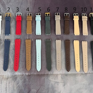 May include: A collection of eleven watch straps in various colors, including white, navy blue, red, brown, light blue, dark green, beige, dark brown, tan, and black. Each strap has a buckle and is displayed on a neutral surface.