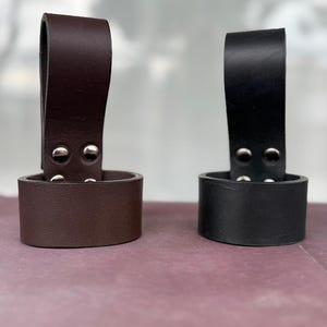 May include: Two leather knife sheaths, one brown and one black, each with a loop handle and secured with silver rivets. The sheaths are cylindrical and designed to hold a knife. The background is a muted purple.