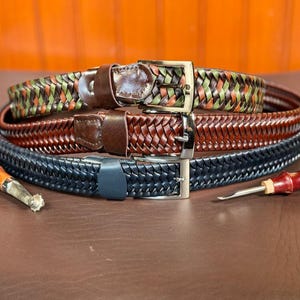 May include: Three braided leather belts in different colors: black, brown, and green. The belts are stacked on top of each other and have silver buckles.