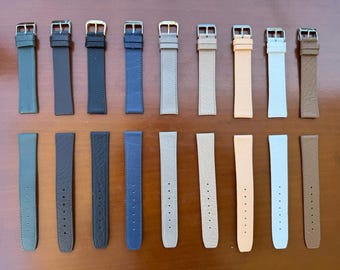 May include: A selection of watch straps in shades of green, black, blue, grey, beige, peach, white, and brown. The leather straps have silver or gold buckles. The straps are displayed in two rows on a wooden surface.
