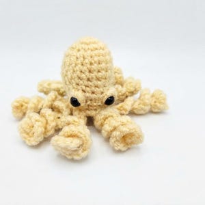 May include: A small, cream-colored crocheted octopus toy featuring a rounded head, eight spiraled tentacles, and two smooth, black plastic safety eyes. The yarn texture is visible throughout the entire handmade stuffed creature.