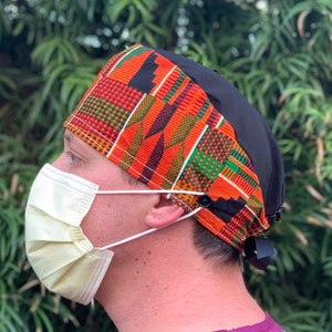 May include: A black and orange patterned surgical scrub hat with a black tie. The hat has a button on the side and is worn by a person wearing a purple scrub top and a yellow face mask.