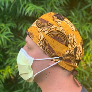 May include: A yellow and brown patterned surgical scrub hat with a black trim and a white tie. The hat is worn by a person wearing a grey scrub top and a light green surgical mask.