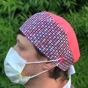 May include: A patterned surgical scrub cap with a pink and white design, and a solid coral pink panel. The cap has a button and white ties. A white face mask is worn with the cap.