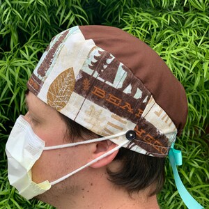 May include: A brown and white patterned scrub hat with a brown top and a turquoise ribbon. The hat has a button on the side and the words "Bear" and "Creek" are visible on the fabric.