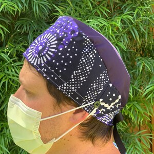 May include: A patterned surgical scrub cap with a purple and black design. The cap features a button and a black tie in the back. The person in the image is wearing a yellow surgical mask.
