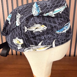 May include: A black scrub cap featuring a repeating pattern of various fish in shades of blue, white, and yellow, set against a dark gray net-like background. The cap includes a small black button on the side and a black ribbon tie at the back.