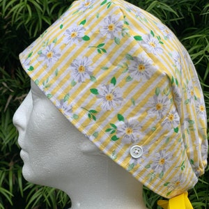 May include: A yellow and white striped scrub hat with a white daisy print and a yellow button. The hat has a yellow tie.