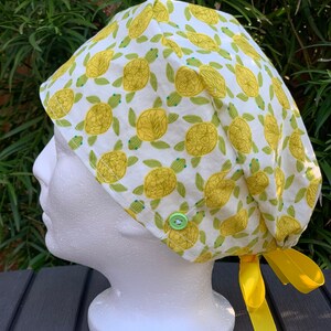 May include: A white scrub hat with a yellow and green turtle print. The hat has a yellow tie and a green button.