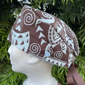 May include: Brown and white patterned surgical scrub hat with a blue button and brown ties. The hat features a Polynesian design.