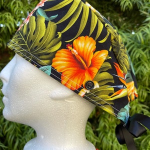 May include: Black scrub hat with a tropical print featuring orange hibiscus flowers, green leaves, and a blue bird. The hat has a button and ties.