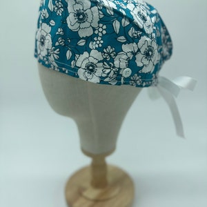 May include: A teal blue surgical scrub hat with a white floral pattern. The hat has a white tie and a button on the side.