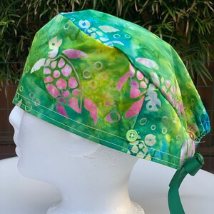 May include: Green, blue, and pink surgical scrub hat with a tie. The hat has a tie-back closure and a button on the side. The fabric has a tie-dye pattern with sea turtles and bubbles.