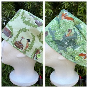 May include: Two green scrub hats with a jungle theme. One hat features a snake, Mowgli, and Baloo. The other hat features King Louie, Shere Khan, and Mowgli. Both hats have green buttons.