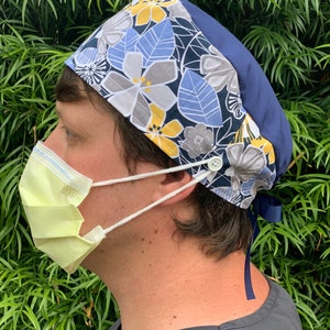 May include: A blue and white floral scrub cap with a yellow trim and a button closure. The cap has a tie at the back.