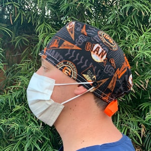 May include: A black and orange scrub hat with a baseball theme. The hat features the San Francisco Giants logo and the words "Giants" and "San Francisco".