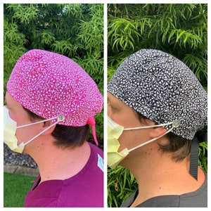 May include: Two surgical scrub caps with a white and pink floral pattern and a white and black floral pattern. The caps have a button and tie closure.