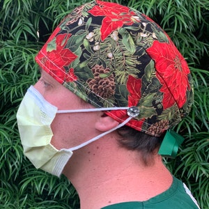 May include: A Christmas-themed surgical cap with a festive poinsettia and pinecone pattern in red, green, and gold on a black background. The cap is paired with a surgical mask and a green scrub top.
