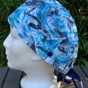 May include: A blue and white surgical scrub hat with a whale and wave pattern. The hat has a blue button and navy blue ties.