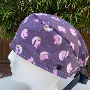 May include: A purple scrub hat with a repeating pattern of pink cartoon pigs on a crescent moon with yellow stars. The hat has a black tie and a black button.