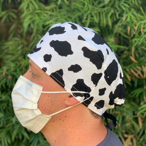 May include: A black and white cow print surgical scrub hat with a black button and tie. The hat is worn by a person wearing a grey shirt and a white face mask.