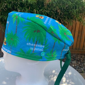 May include: A blue surgical scrub cap with a tropical palm tree pattern and the text "cheeseburger in paradise." The cap has a green trim, a green button, and green ties.