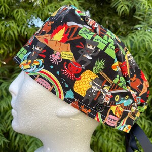 May include: A black scrub hat with a colorful Hawaiian-themed print featuring cartoon characters, pineapples, and text that says "Kilauea Visitor Center", "Lani", "Plantation Garden", and "Waimea Bay Beach".