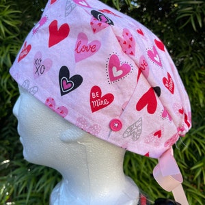 May include: A pink scrub cap with a pattern of red, pink, and black hearts, with the words "love", "xo", and "be mine". The cap has a pink ribbon and a pink button.