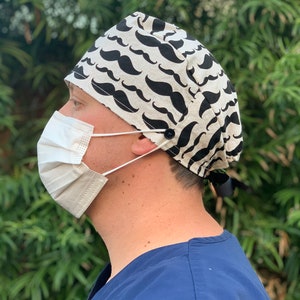 May include: A white and black patterned scrub hat with a black mustache design. The hat has a button and tie closure.