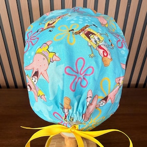 May include: A light blue scrub cap featuring a repeating pattern of SpongeBob SquarePants and Patrick Star characters, accented with pink flower shapes and yellow bubbles. The cap includes a bright yellow ribbon tie at the back for adjustment.