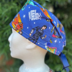 May include: Blue surgical scrub hat with a print of characters from the Big Bang Theory television show. The hat has a button closure and a navy blue tie.