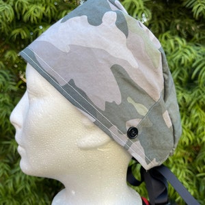 May include: A camouflage patterned surgical scrub hat with a black button and black ties. The hat is on a white mannequin head.
