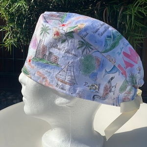 May include: A white scrub cap with a Hawaiian-themed print featuring illustrations of palm trees, sailboats, and landmarks. The cap includes text such as "Yellow Hibiscus" and "Maui". A cream-colored ribbon is attached to the back of the cap.
