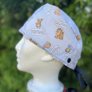 May include: A light blue surgical scrub cap featuring Garfield comic strip illustrations. The cap has a black button and black ties. Text includes phrases like "I have to break this teddy bear dependency."