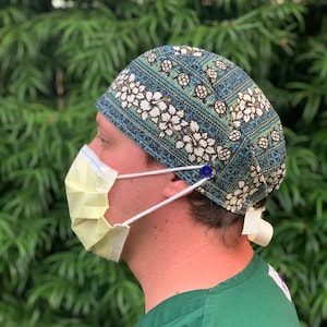 May include: A patterned surgical scrub cap with white floral designs on a teal background. The cap is secured with a blue button and a white ribbon tie. A yellow face mask is worn with the cap.