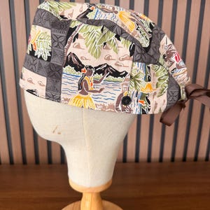 May include: A scrub cap featuring a tropical print with hula dancers, palm trees, and mountains in shades of tan, green, and black. The fabric includes geometric patterns, a small black button on the side, and a brown ribbon tie at the back.