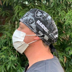 May include: A black and white surgical scrub hat with a repeated pattern of the Las Vegas Raiders logo and the word "Raiders".