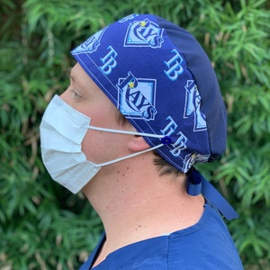 May include: A blue and white surgical scrub hat with a repeating pattern of the Tampa Bay Rays logo and the letters "TB". The hat has a black back and a white adjustable tie.